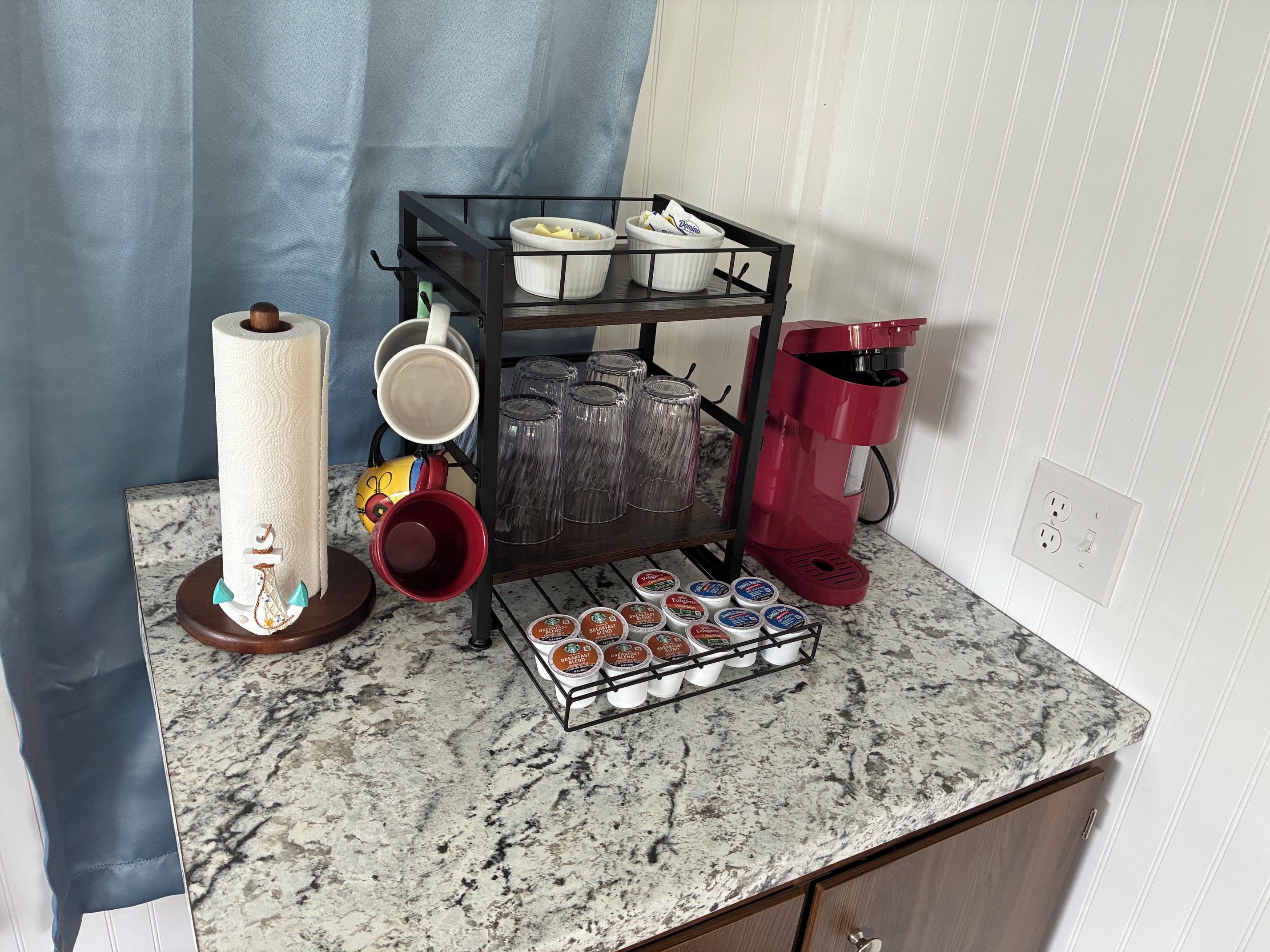 Una encimera de cocina con una cafetera, un escurreplatos para tazas, un rollo de toallas de papel y una bandeja de cápsulas de café.