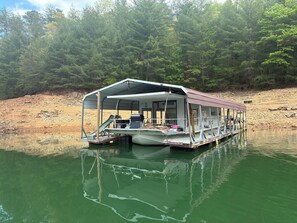 A floating houseboat with a covered deck, a grill, and a small boat docked at the front.
