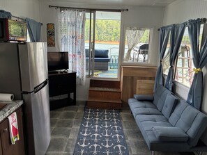 A compact living space with a kitchenette, a sofa, a television, and a view of a dock and water.