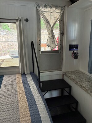 A room with a bed, a window with a curtain, a small table, and a wall-mounted speaker.