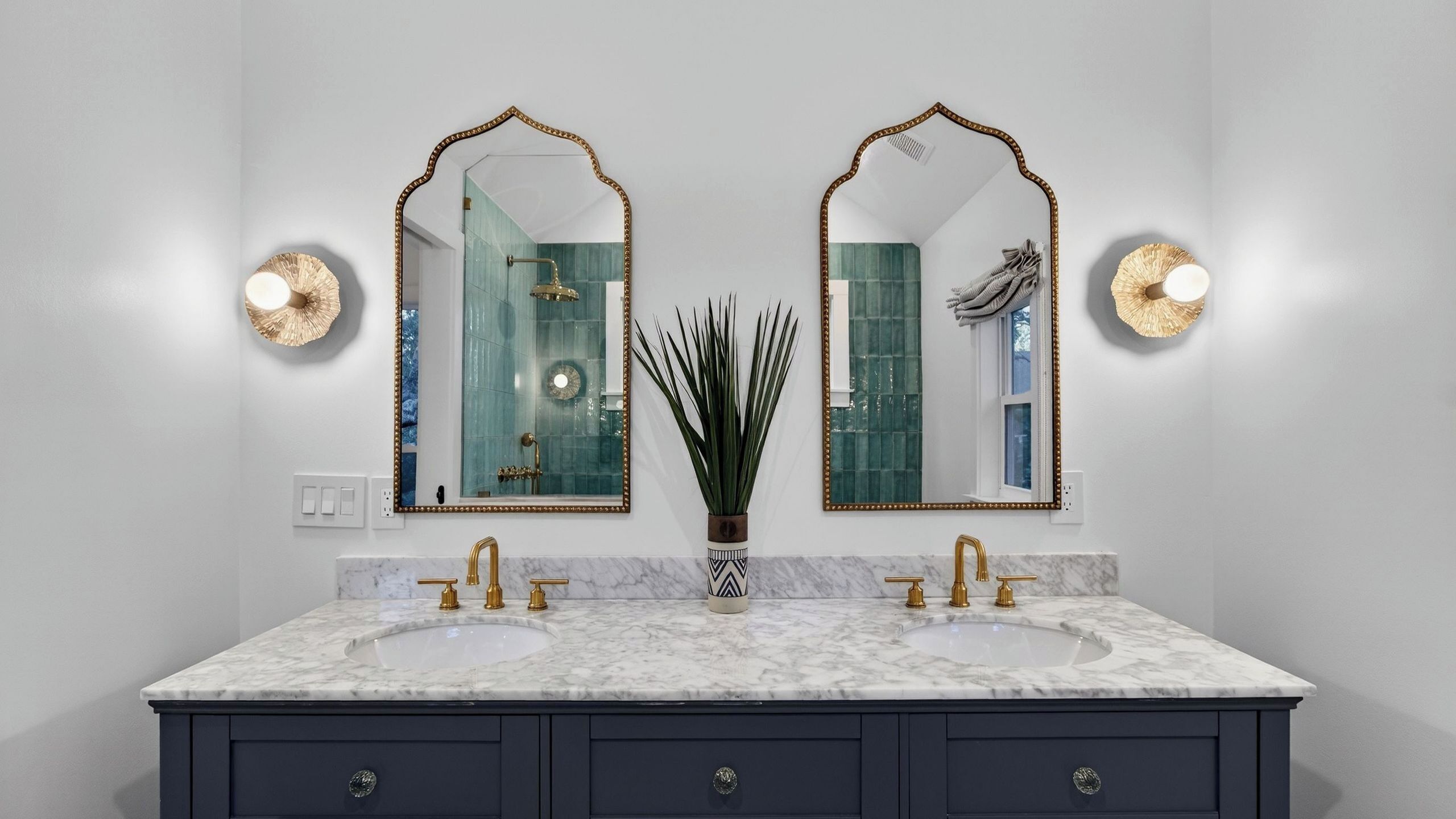 A bathroom with two mirrors, a marble sink, and a vase with tall plants.