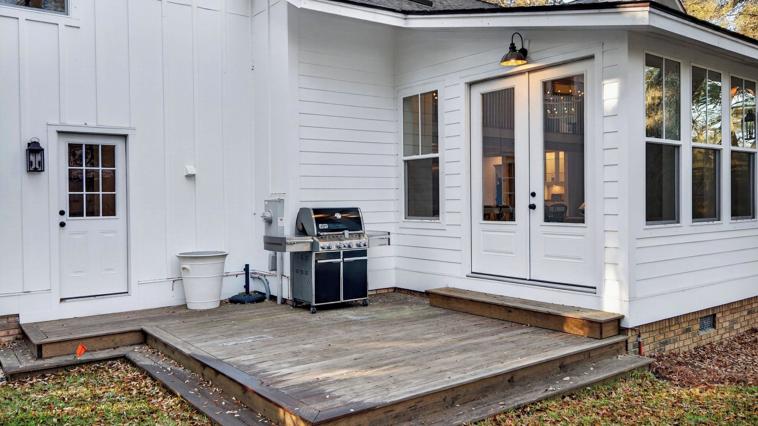 Een wit huis met een overdekte veranda, een barbecue en een vuilnisbak.