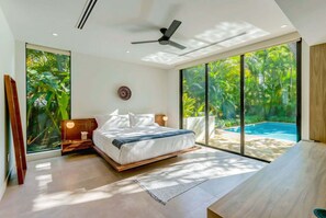 Quarto moderno com cama, ventilador de teto, mesa de cabeceira e grande janela com vista para uma piscina.