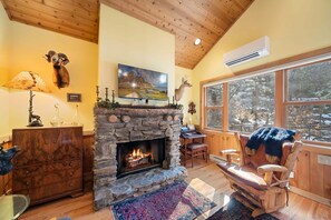 A cozy room with a stone fireplace, wooden furniture, and a large window with a view of trees. - Charming Beech Mtn Retreat Near Slopes | Cozy Fireplace Escape (Banner Elk)