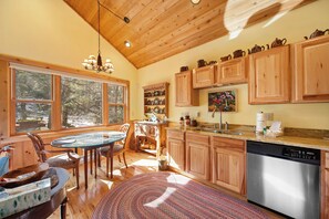 A wooden interior with a dining table, chairs, a kitchen with wooden cabinets, a dishwasher, and a large window. - Charming Beech Mtn Retreat Near Slopes | Cozy Fireplace Escape (Banner Elk)