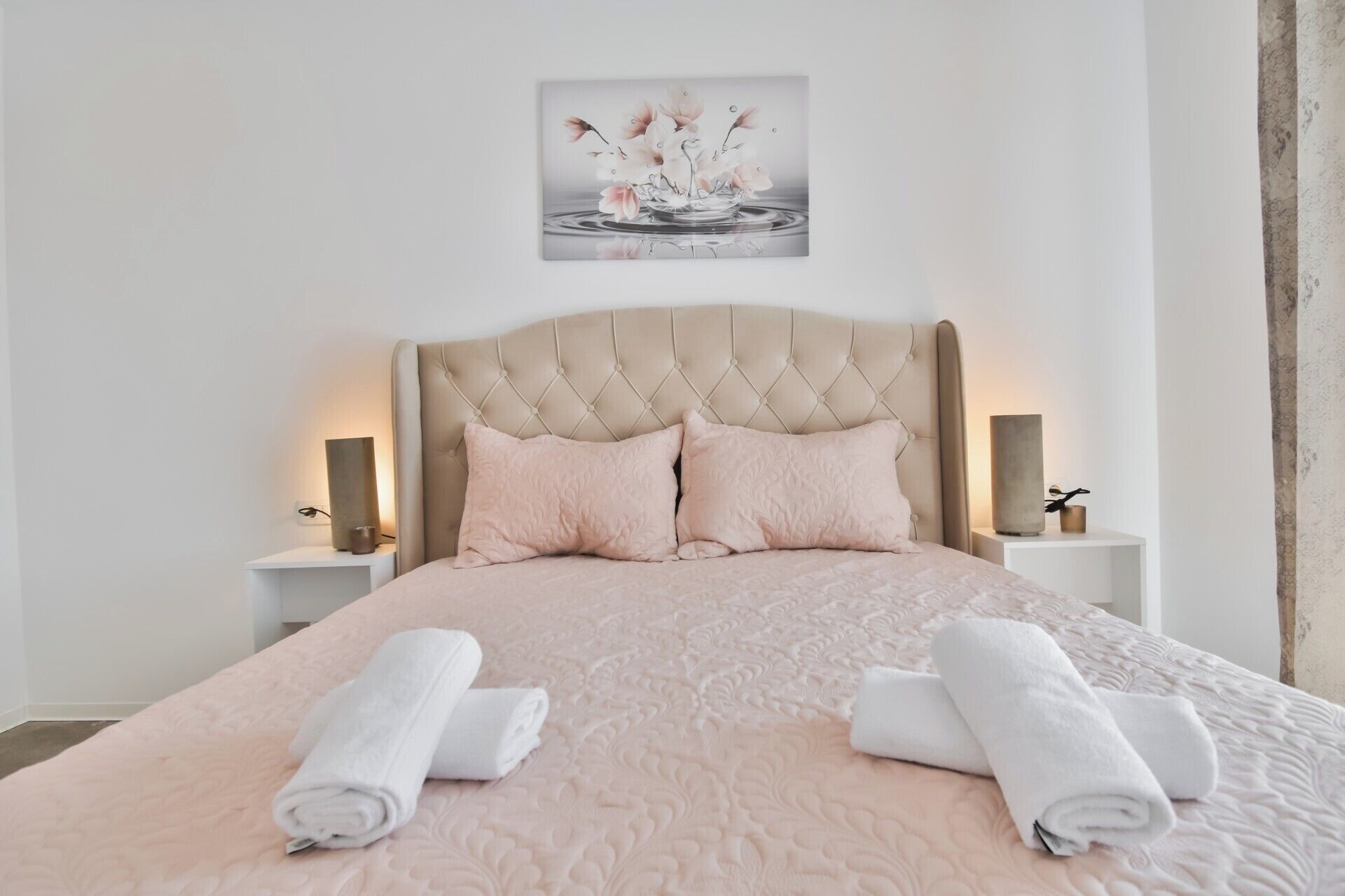A neatly made bed with a pink quilt, two pillows, and rolled towels on a white headboard.