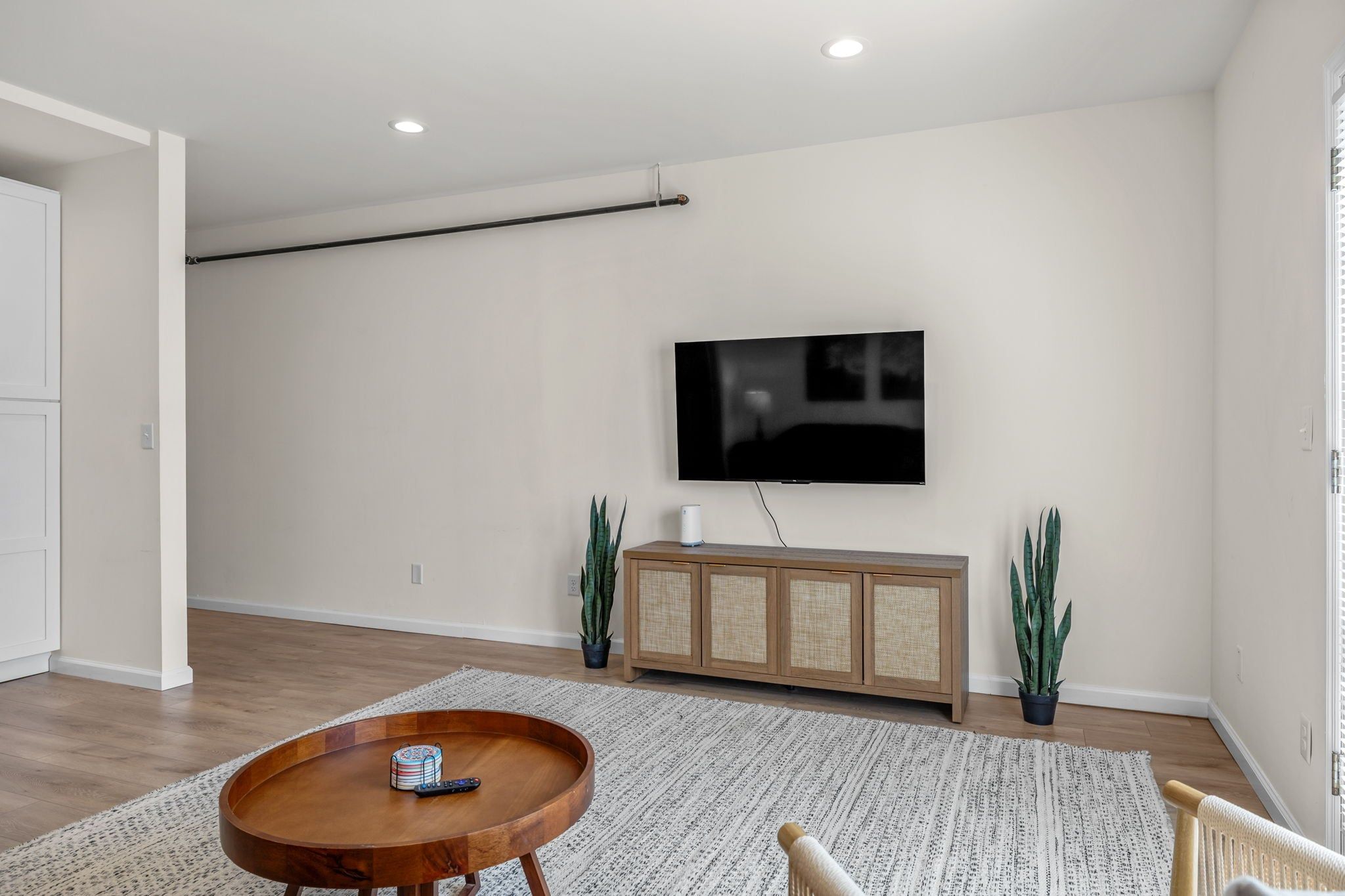 Una sala de estar moderna con un televisor de pantalla plana montado en la pared, una mesa de comedor de madera con un control remoto, un tapete estampado y dos plantas en macetas.