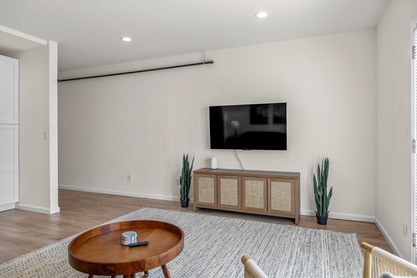 A modern living room with a flat-screen TV mounted on the wall, a wooden dining table with a remote control, a patterned rug, and two potted plants.