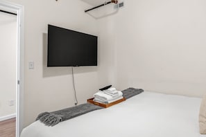 A hotel room with a bed, a flat-screen TV mounted on the wall, a wooden bedside table with a towel and a remote control, and a grey throw blanket.
