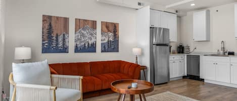 A modern living room with a sofa, a round wooden coffee table, and a chair. There are three wall-mounted paintings, a kitchen area with a refrigerator, and a sink.