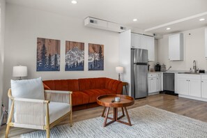A modern living room with a sofa, a round wooden coffee table, and a chair. There are three wall-mounted paintings, a kitchen area with a refrigerator, and a sink.