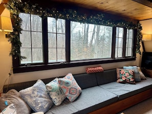 Ein gemütliches Zimmer mit einem großen Fenster, einer Couch mit gemusterten Kissen und einem Fernseher auf einem Holztisch.