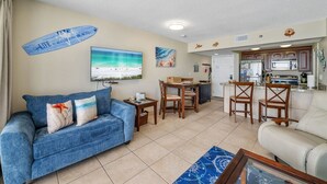 Una sala de estar con un sofá azul, un comedor con sillas de madera, una cocina con armarios oscuros y un televisor grande que muestra una escena de playa.
