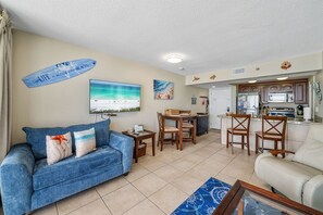 Ein Wohnzimmer mit blauem Sofa, einem Essbereich mit Holzsesseln, einer Küche mit dunklen Schränken und einem großen Fernseher, der eine Strandansicht zeigt.