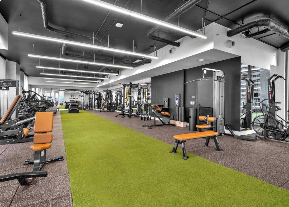 A gym with exercise equipment, green artificial turf, and a row of orange benches.
