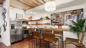 A modern kitchen with a stainless steel refrigerator, a microwave, and a wooden ceiling. There are bar stools and a bar counter. - Contemporary 3BR Condo | Downtown Near Nightlife (Savannah)