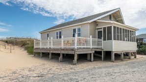 Une maison située en bord de mer, dotée d’une terrasse en bois, d’un porche abrité et d’une plage de sable en arrière-plan.
