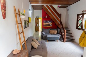 A living room with a sofa, a wooden ladder, a staircase, and a red wall with a painting. - Casa com 2 suítes e Churrasqueira no Figueira (Ubatuba)