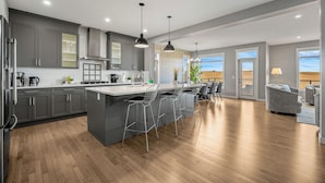 A modern kitchen with grey cabinets, a central island, bar stools, and a dining area with a grey sofa.