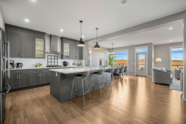 A modern kitchen with grey cabinets, a central island, bar stools, and a dining area with a grey sofa.