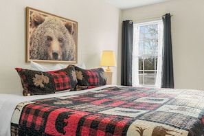 Une chambre à coucher avec un grand lit, une couette, un oreiller, une lampe, une fenêtre avec des rideaux et un tableau représentant un ours accroché au mur.