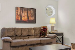 Una sala de estar con un sofá color crema, un cuadro de un bosque enmarcado, una lámpara y una mesita.
