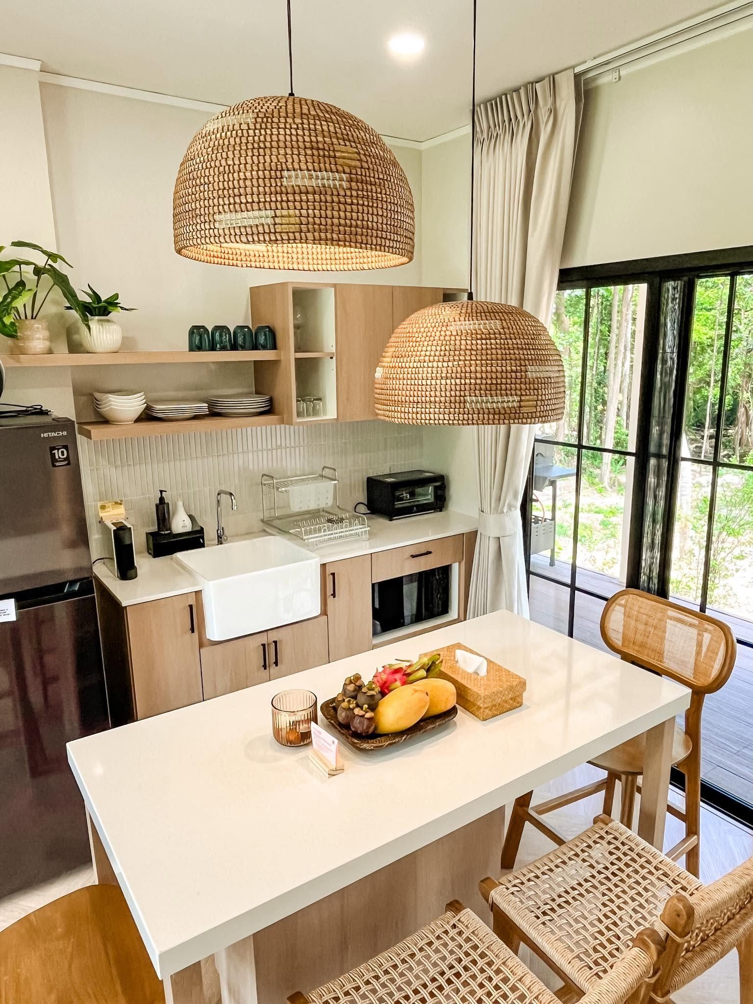 A modern kitchen with a central island, pendant lights, and a view of greenery outside.