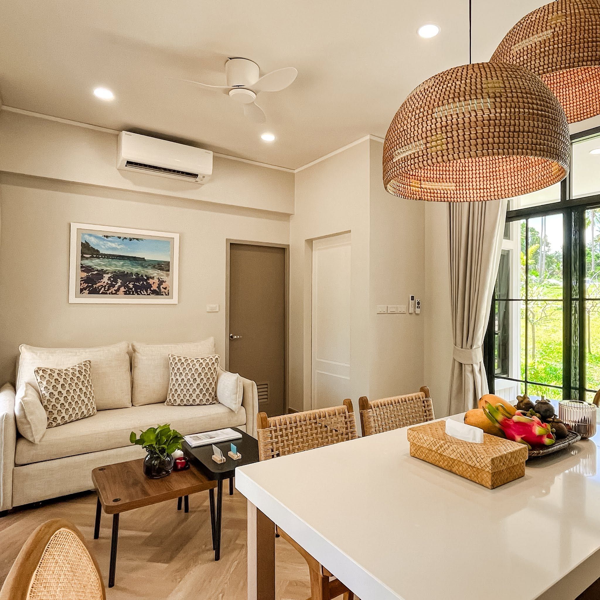 Un salon moderne comprenant un canapé, une table basse, une table à manger, des fauteuils en osier et une grande fenêtre donnant sur un paysage verdoyant.