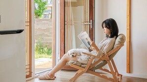 A person reading a book in a room with a wooden floor, a sliding glass door leading to an outdoor area, and a small white dog resting on their lap.