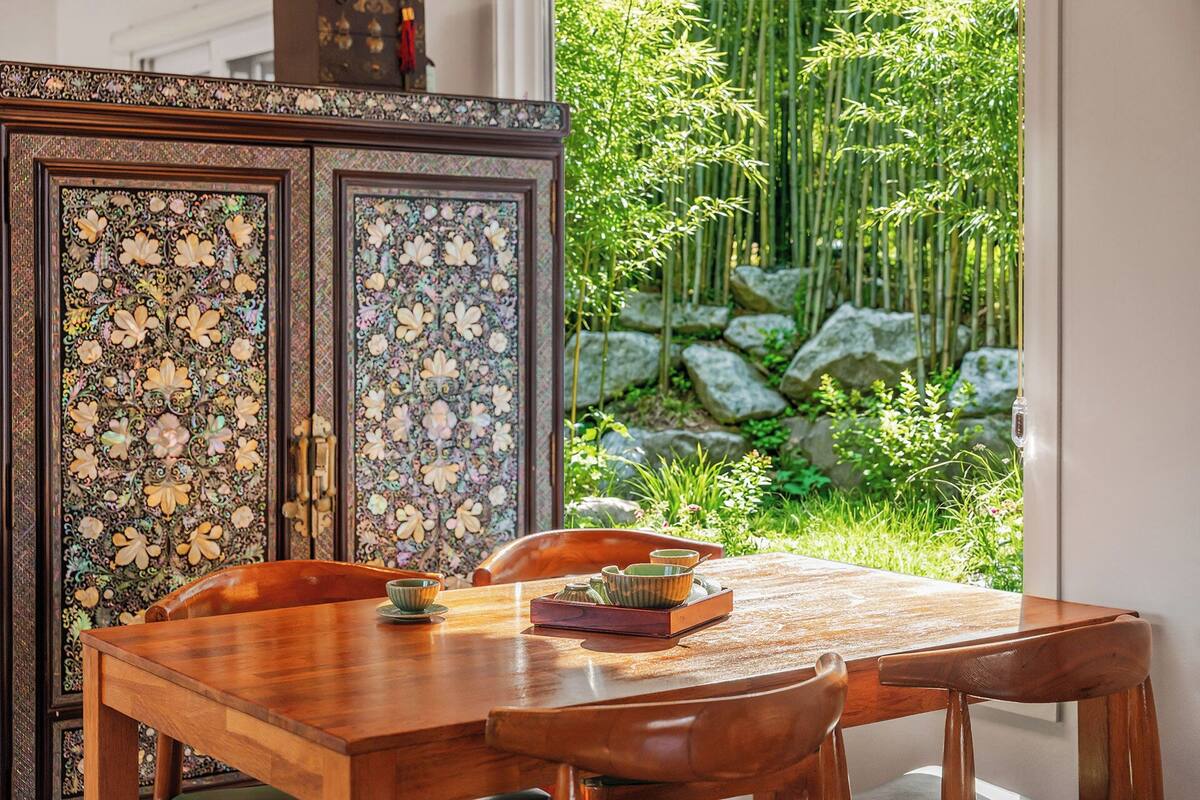 Un comedor con una mesa y sillas de madera, un mueble decorativo con motivos florales y vistas a un jardín de bambú.