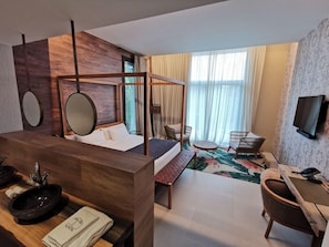 Ein Hotelzimmer mit Schreibtisch aus Holz, einem Himmelbett, einem an der Wand montierten Fernseher und einem großen Fenster mit Vorhängen.