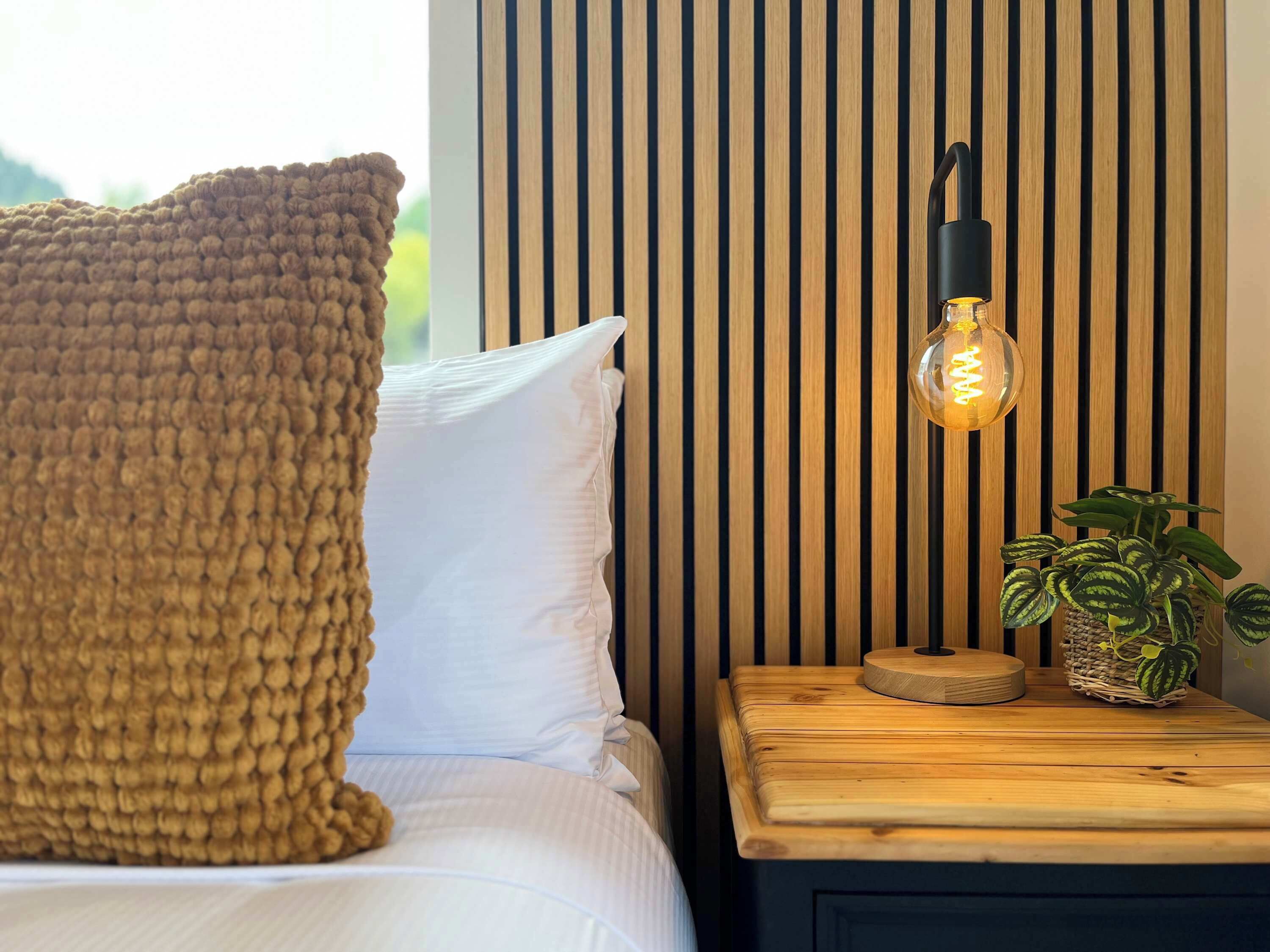 A bed with white bedding, a brown knitted throw, a white pillow, a wooden nightstand with a lamp, and a potted plant.
