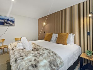 A modern hotel room with a large bed, a wooden slat wall, a mounted TV, and a small side table with a plant. - Smart Studio Stay | Great Value Location (Queenstown)