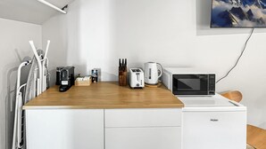 Una cocina compacta con encimera de madera, armarios blancos, microondas, tostadora, cafetera y un juego de cuchillos. Un televisor montado en la pared muestra un paisaje montañoso.