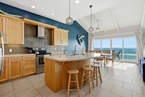 A kitchen with wooden cabinets, a large island with stools, a stove, and a view of the ocean through large windows. - Pacific Edge Retreat | Entire Estate, Ocean Views, Luxury Coastal Escape (Oceanside)