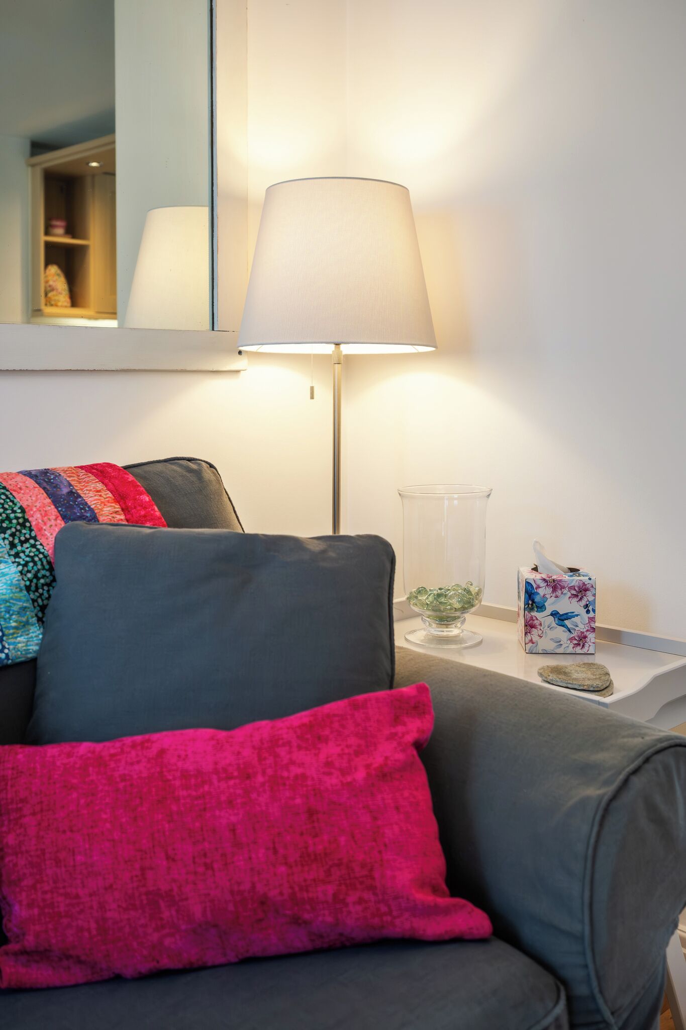 Una sala de estar moderna con un sofá gris, un cojín rosa, una lámpara de pie, un florero de vidrio y una caja de pañuelos.