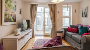 A living room with a sofa, a television, a coffee table, and a large window with curtains. - Beautiful Two BD Home Near Cirencester (Stratton)