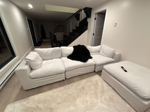 Un canapé d’angle blanc, recouvert d’un plaid noir, avec un pouf blanc et une télécommande posée dessus.