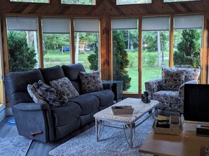 Ein Zimmer mit einem dunkelgrauen Sofa, einem gemusterten Sessel, einem kleinen Tisch und einem Schreibtisch mit Monitor und Tastatur.