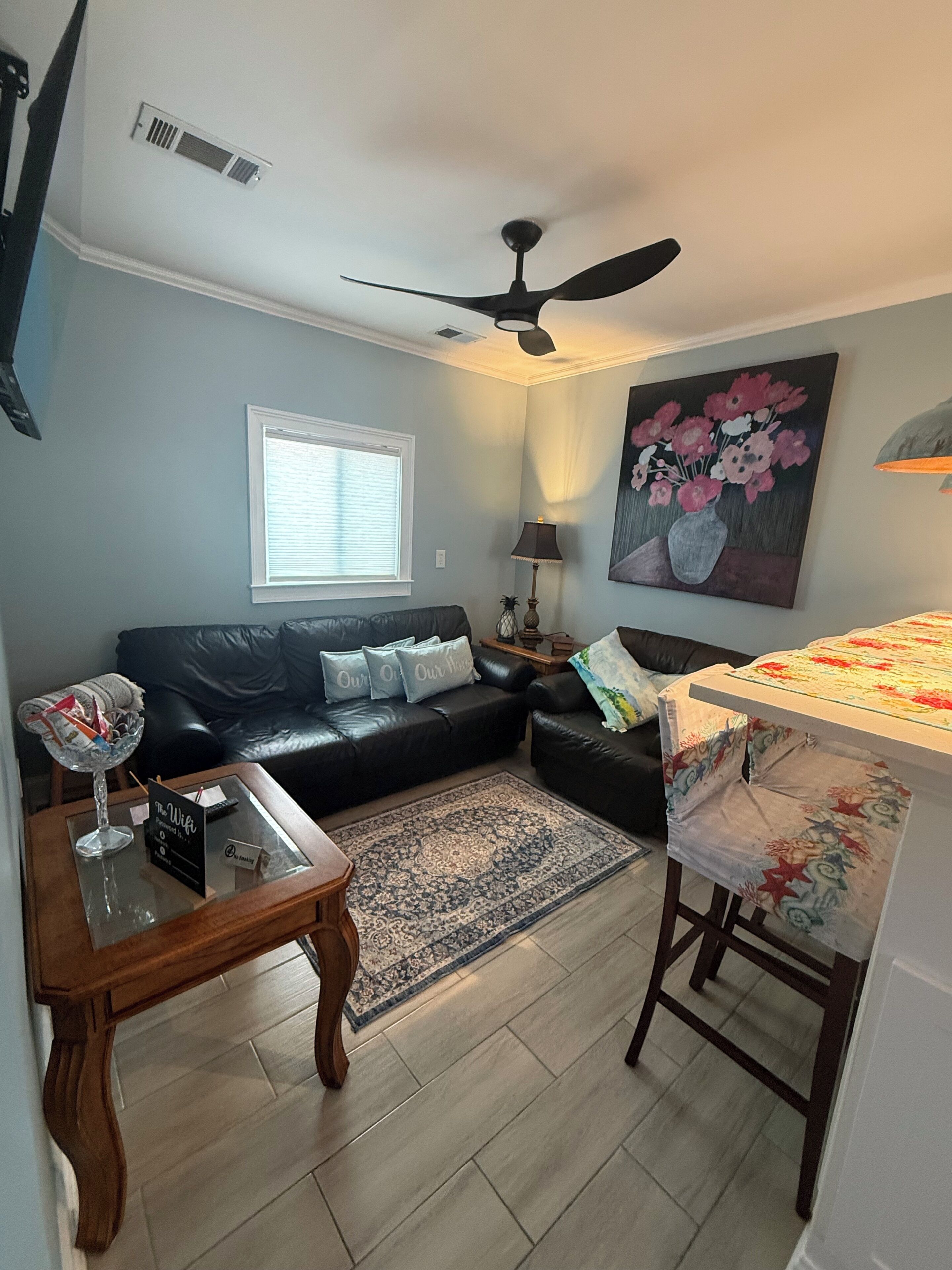A living room with a black leather sofa, a glass coffee table, a wooden side table, a patterned rug, a ceiling fan, a painting, and a window with blinds.