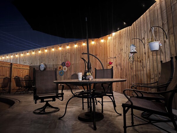 An outdoor patio with a round table and chairs, a wooden wall, and string lights.