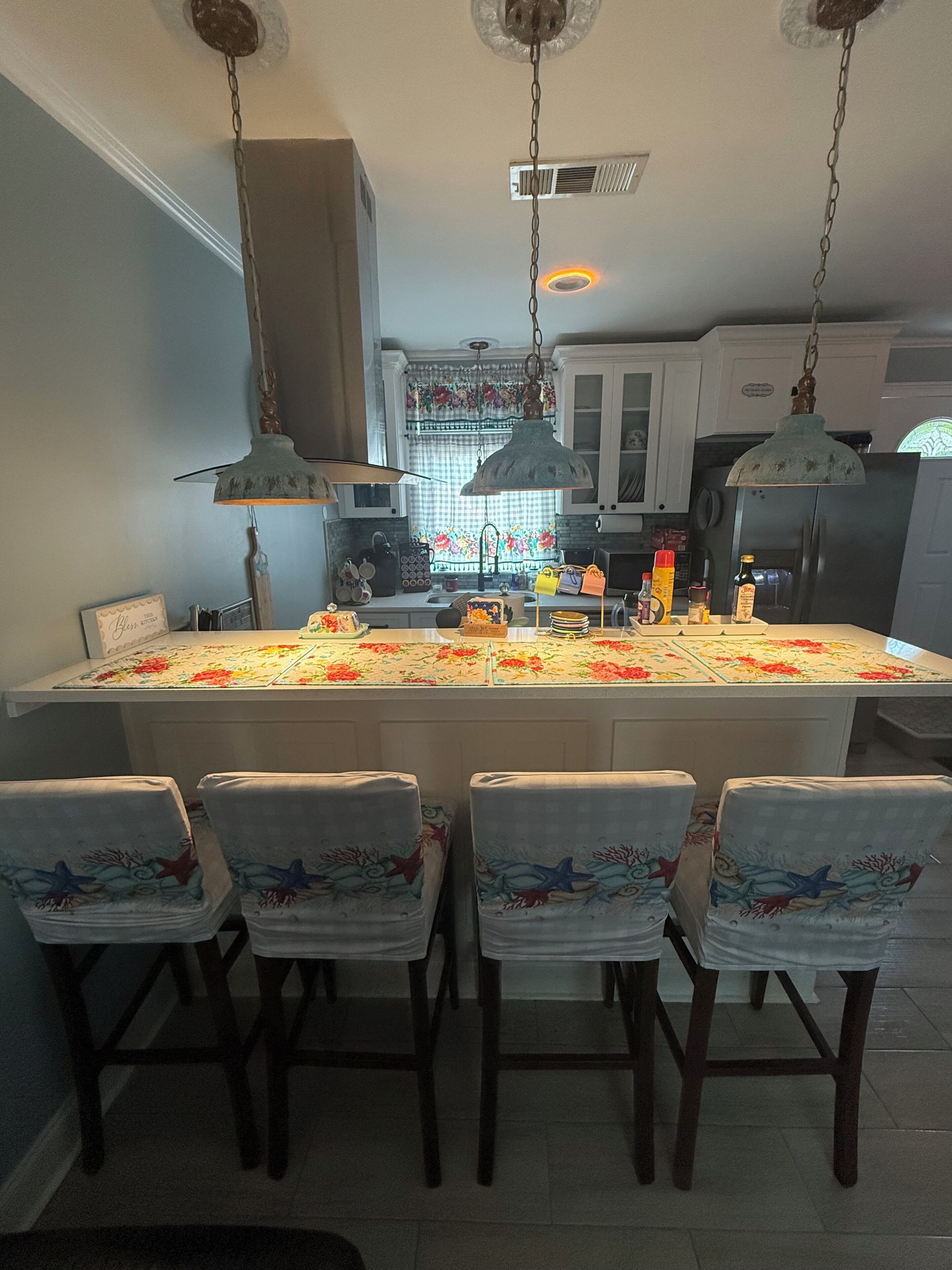 Een keuken met een centraal eiland, stoelen met bloemenmotieven, hanglampen en een koelkast.