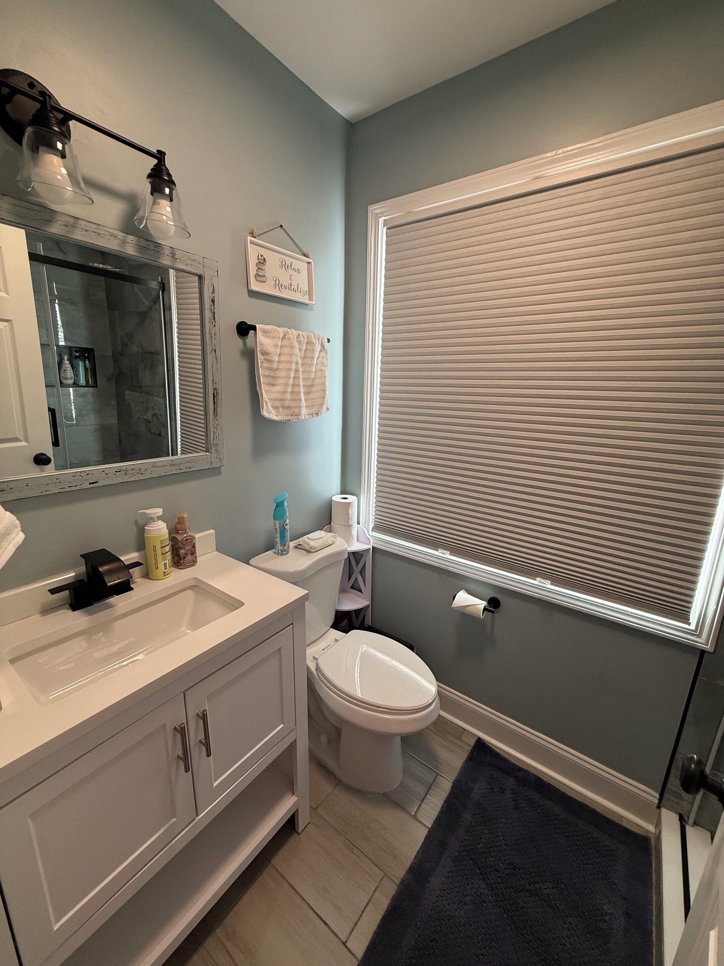 A bathroom with a white vanity, a mirror, a toilet, and a window with blinds.