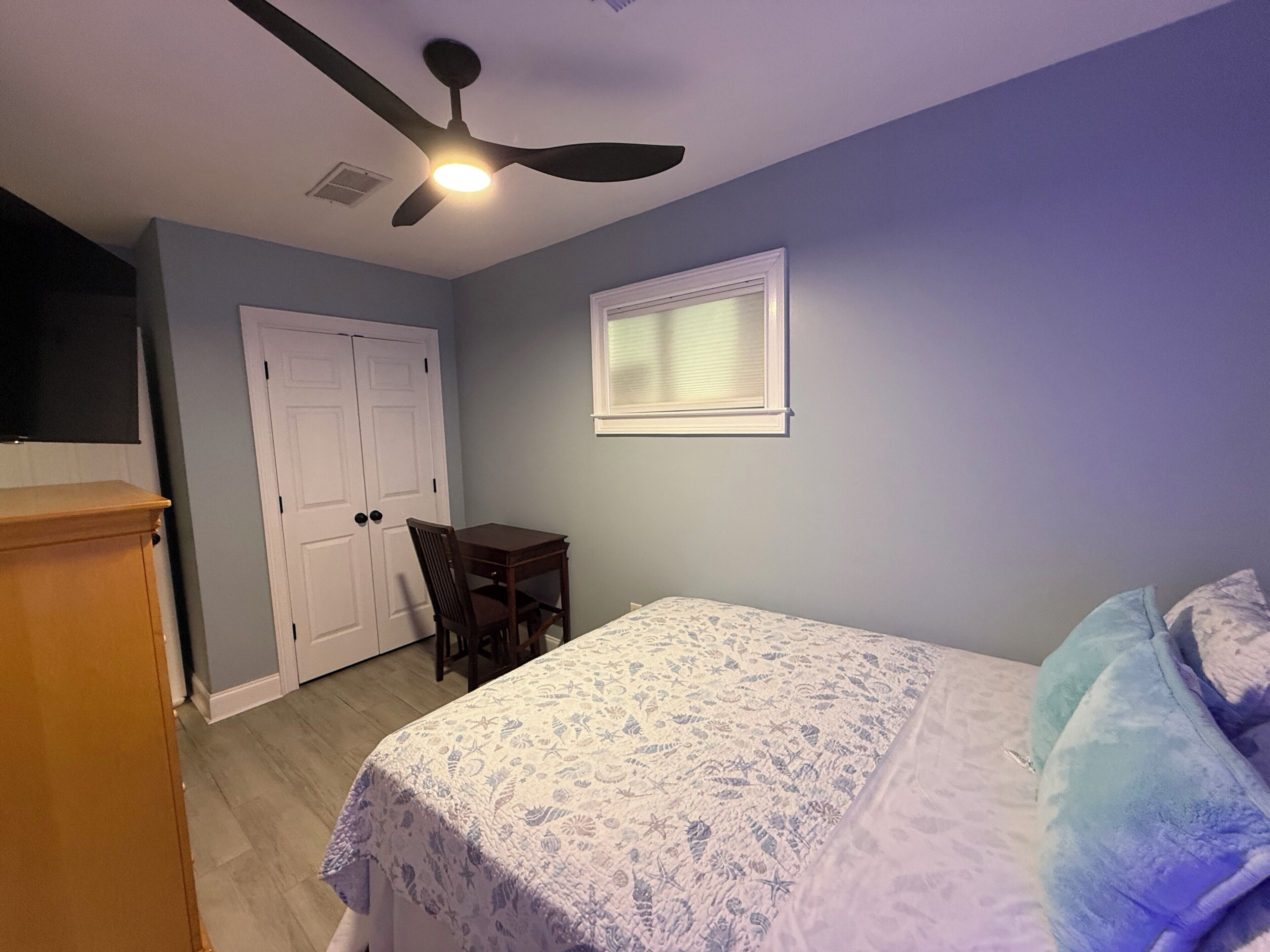 A bedroom with a bed, a desk, a chair, a ceiling fan, and a window.