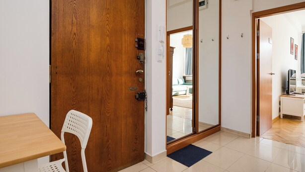 A wooden door, a white chair, a wooden table, and a glimpse of a living area with a sofa and a TV. - Plac Wilsona Charming Apartment (Warszawa)