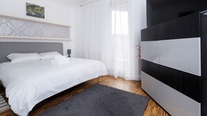 Une chambre à coucher moderne avec un lit, un parquet en bois foncé, un tapis gris, une tête de lit blanche, une armoire noire et blanche, et une fenêtre avec des rideaux légers.