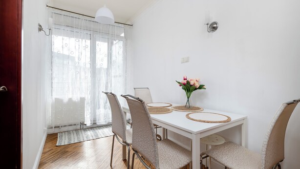 Un coin repas avec une table blanche, quatre chaises, un vase de fleurs et une fenêtre ornée de rideaux légers.