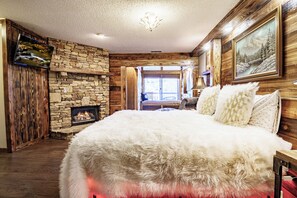 A room with a stone fireplace, a large bed with white bedding, wooden walls, and a chandelier. - Luxury 6th Floor Penthouse • Cabin Vibes + Space Needle & Downtown Views (Gatlinburg)
