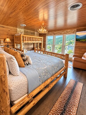 Ein Schlafzimmer einer Holzhütte mit einem großen Bett, einem Holzkopfstück, einer Chandelieber und Blick ins Freie.