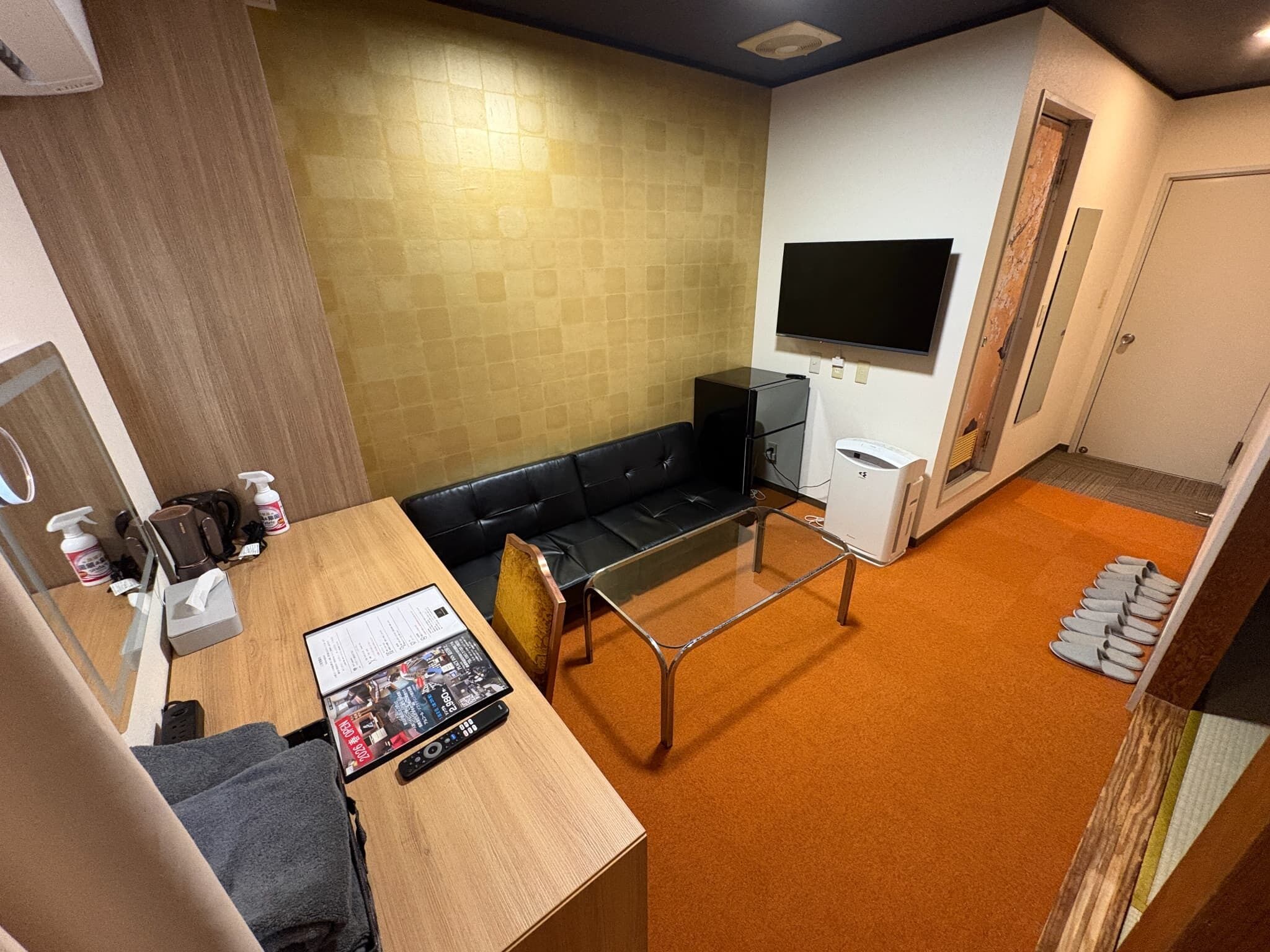 A hotel room with a wooden desk, a black leather sofa, a flat-screen TV mounted on the wall, and a small kitchenette area.